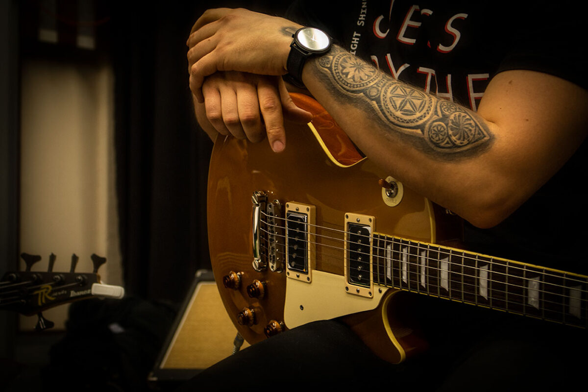 Kitaristi nojaa kitaraansa. Kuvassa näkyy vain kitaristin tatuoidut kädet ja kello.
