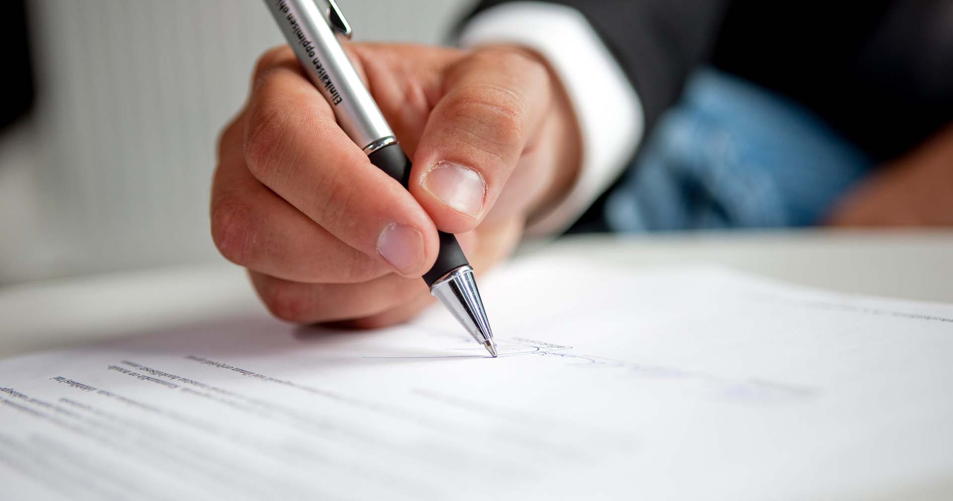 Lähikuva kädestä, joka pitää kynää ja allekirjoittaa asiakirjan pöydällä, paperilla näkyy tekstiä. Henkilöllä on yllään puvun takki.