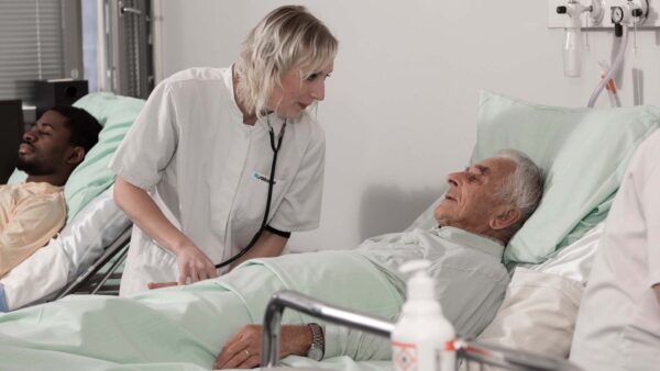 Sairaanhoitaja on sairaalahuoneessa ikääntyneen potilaan äärellä. Potilas makaa sairaalasängyssä selällään.