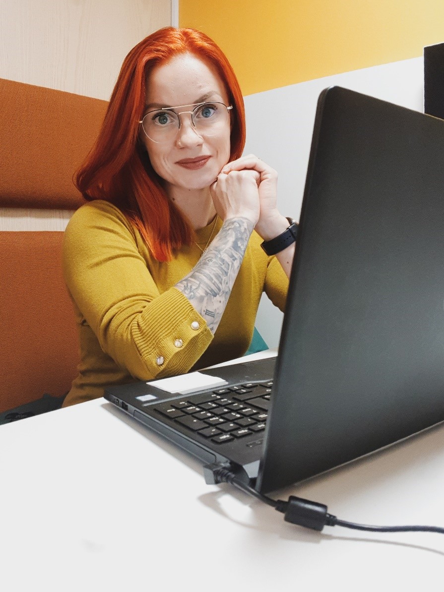 Punatukkainen ja silmälasipäinen nainen istumassa kannettavan tietokoneen ääressä.