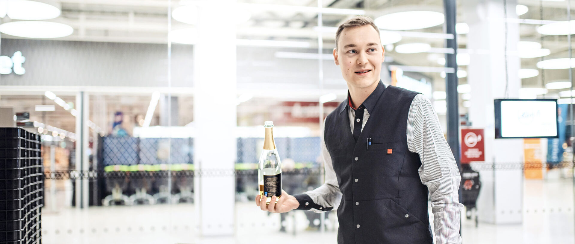 Markus Niskanen työpaikallaan Alkossa, seisoo sisäänkäynnin luona viinipullo kädessään hymyssä suin.