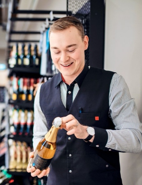 Markus Niskanen työpaikallaan Alkossa, avaamassa viinipulloa hymyssä suin.