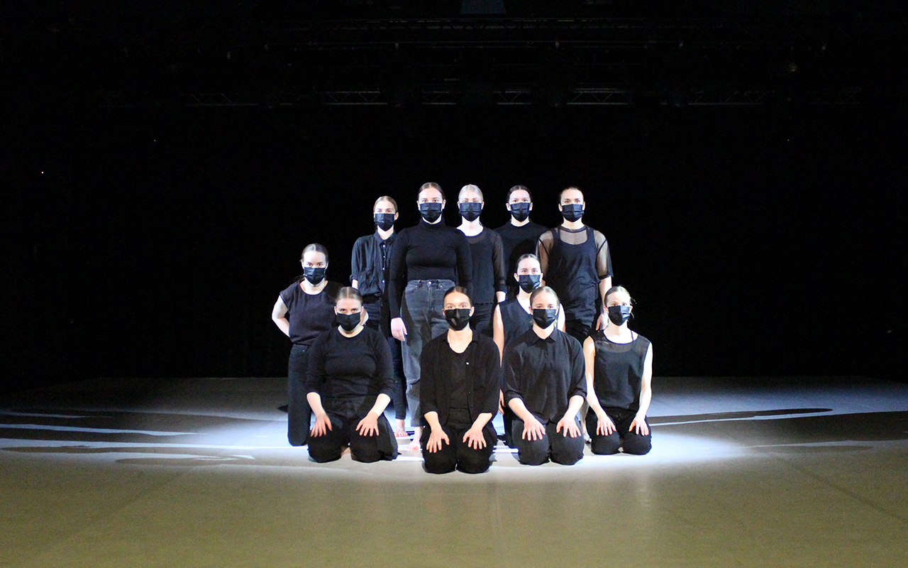 Tanssijat istuvat tai seisovat yhdessä rykelmässä maskit kasvoillaan, valo tekee kuvasta dramaattisen ja sen sävyt ovat hyvin mustavalkoiset.