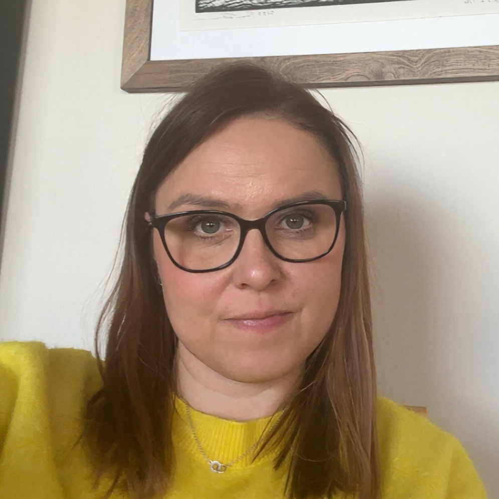 Nainen, jolla on suorat ruskeat hiukset ja mustat silmälasit, on pukeutunut keltaiseen villapaitaan ja katsoo kameraan. Hän istuu sisätiloissa, ja hänen takanaan seinällä on kehystetty kuva.