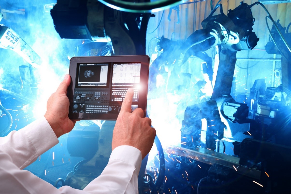 Kuvituskuva. Henkilön kädet näkyvät tablet-tietokoneen päällä ja tabletin taustalla kipinät räiskyvät sinisen sävyisessä konehuoneessa, kun hitsausrobotit työskentelevät.