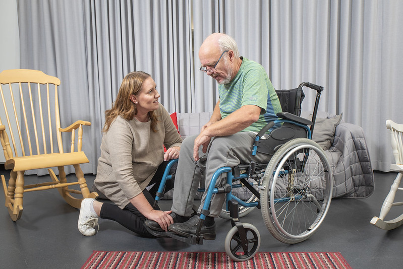 Kuvituskuvassa iäkäs mies istuu pyörätuolissa ja hänen luokseen on kumartunut nuorempi nainen, joka hoitaa häntä.