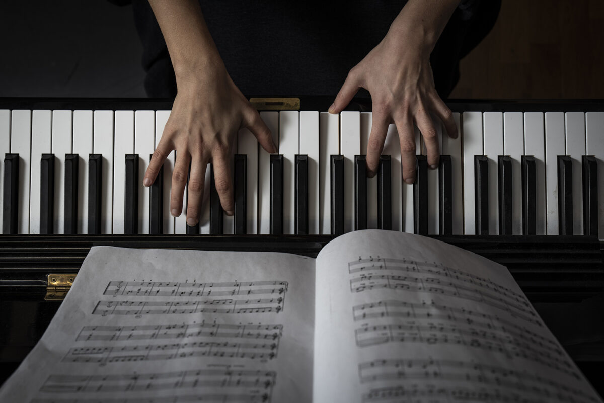 Kuvituskuva. Pianon koskettmien päällä näkyvät sormet ja kuvan alalaidassa nuottivihko, joka on auki soittajalle.