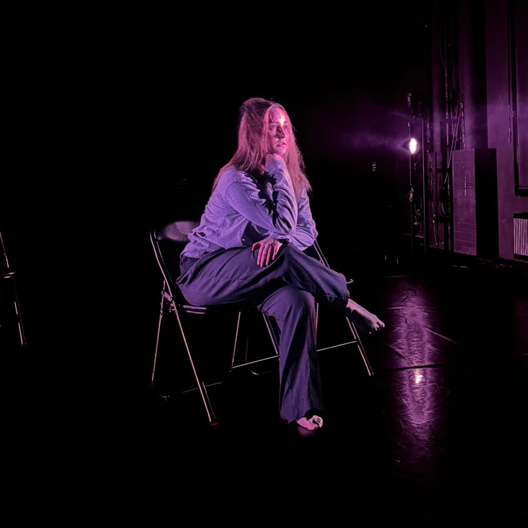 Pitkätukkainen henkilö istuu paljain jaloin taittotuolilla pimeässä huoneessa, jota valaisee dramaattinen vaaleanpunainen ja violetti valaistus, ja hänellä on mietteliäs ilme ja toinen käsi leuan päällä.