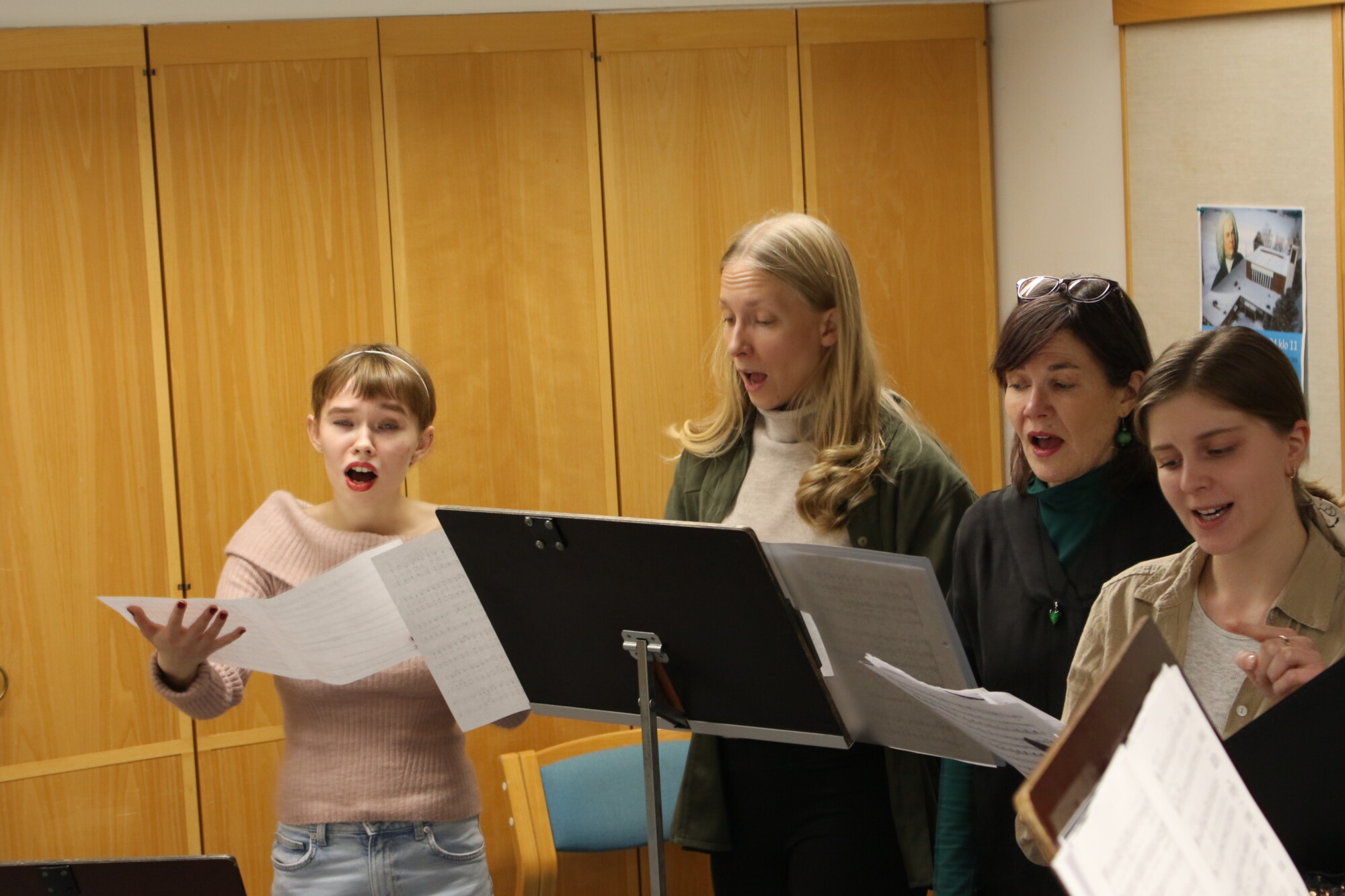 Neljä ihmistä seisoo nuottipöytien takana ja laulaa yhdessä huoneessa, jonka seinät ovat puupaneeleita, pitäen nuotteja käsissään ja vaikuttaen keskittyneiltä esitykseensä.