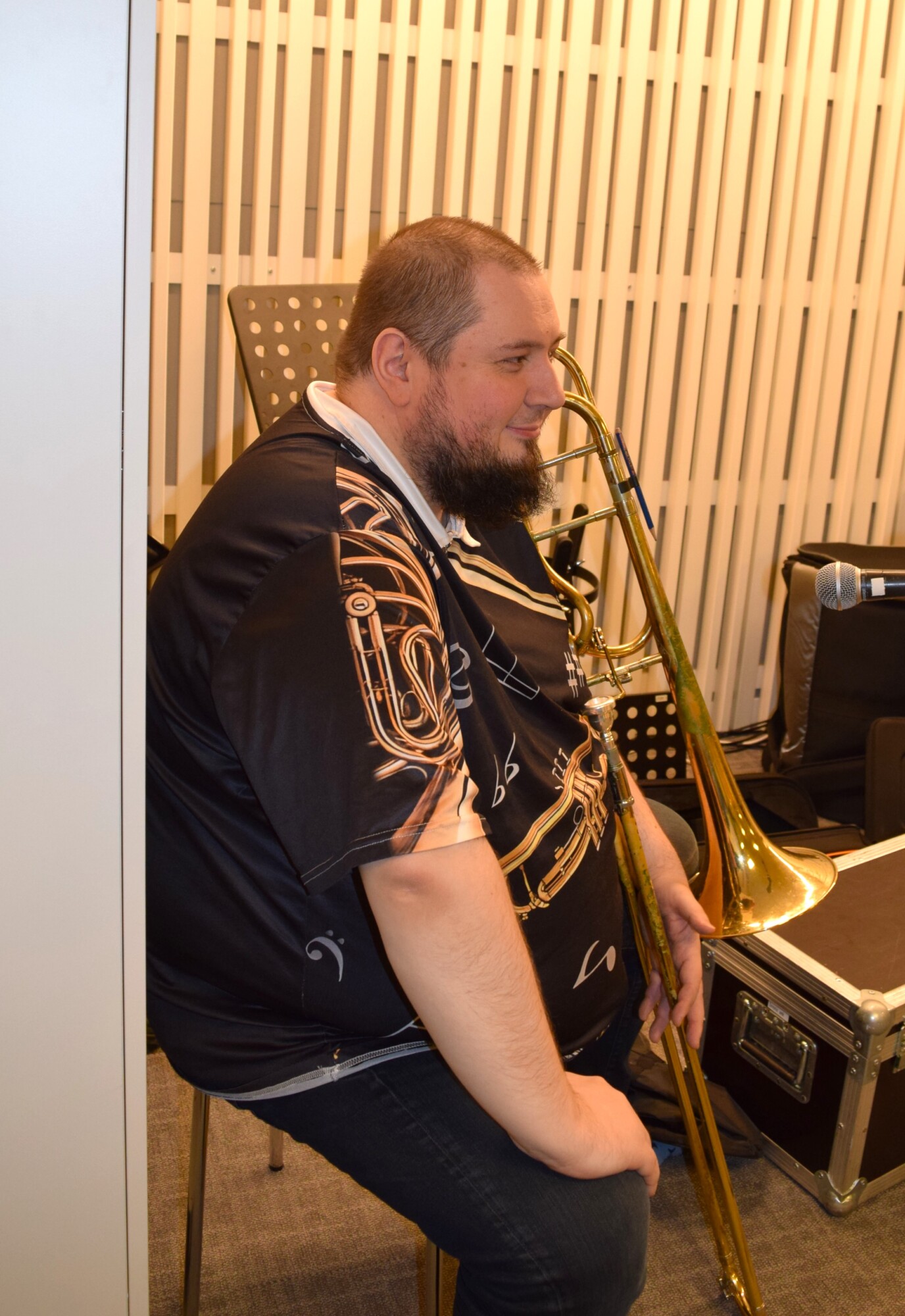 Istuva parrakas mies, jolla on pasuuna kädessään ja yllään musta paita, jossa on nuotteja ja vaskisoittimien kuvioita, istuu huoneessa, jossa on pystysuuntaisia valkoisia seinäpaneeleita ja nuottitelineitä.