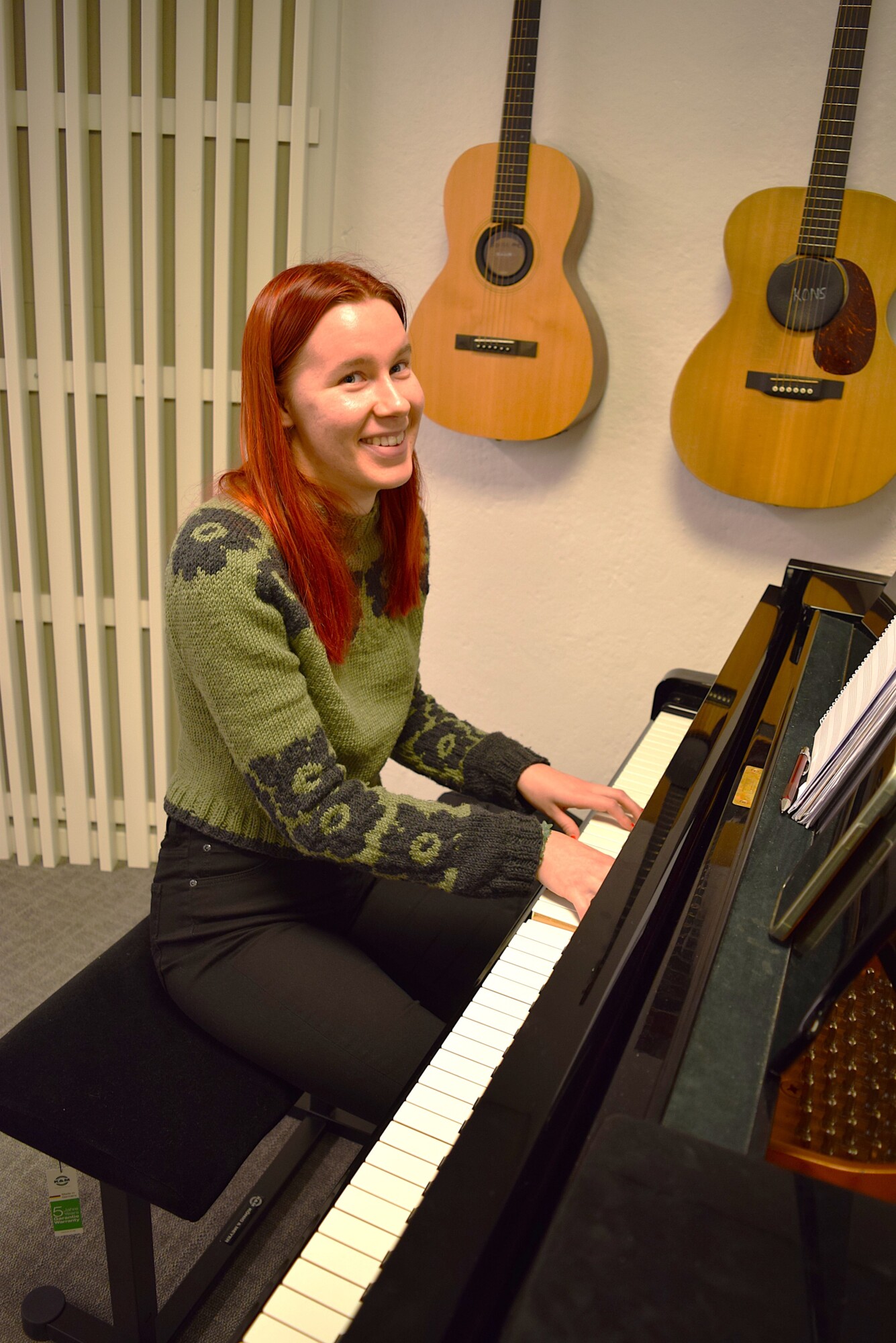 Punatukkainen nainen, jolla on vihreä villapaita, hymyilee soittaessaan pianoa huoneessa, jonka takana seinällä roikkuu kaksi akustista kitaraa.