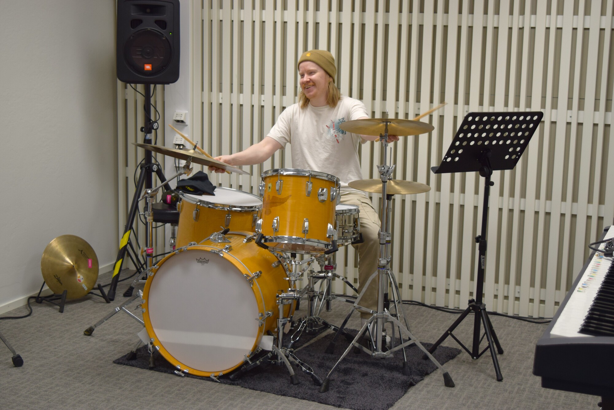 Beigeen pipoon ja valkoiseen paitaan pukeutunut henkilö soittaa keltaista rumpua musiikkihuoneessa. Lähistöllä on symbaaleja, nuottijalusta, näppäimistö ja kaiutin, ja takana seinällä on pystysuorat valkoiset paneelit.