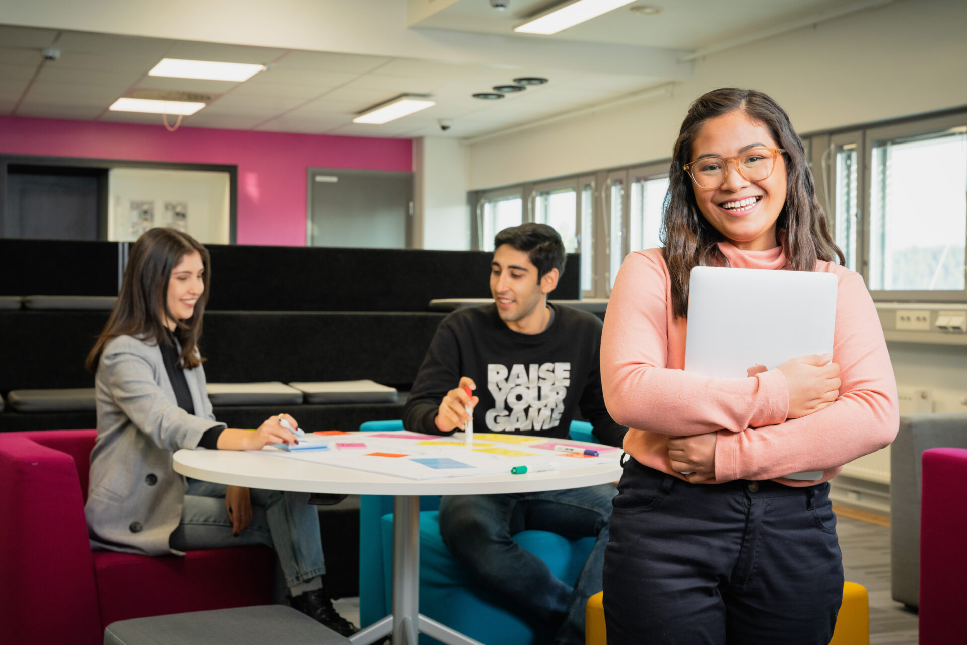 A picture of a student holding a computer in her arms. In the background there are two more students sitting down and discussing.