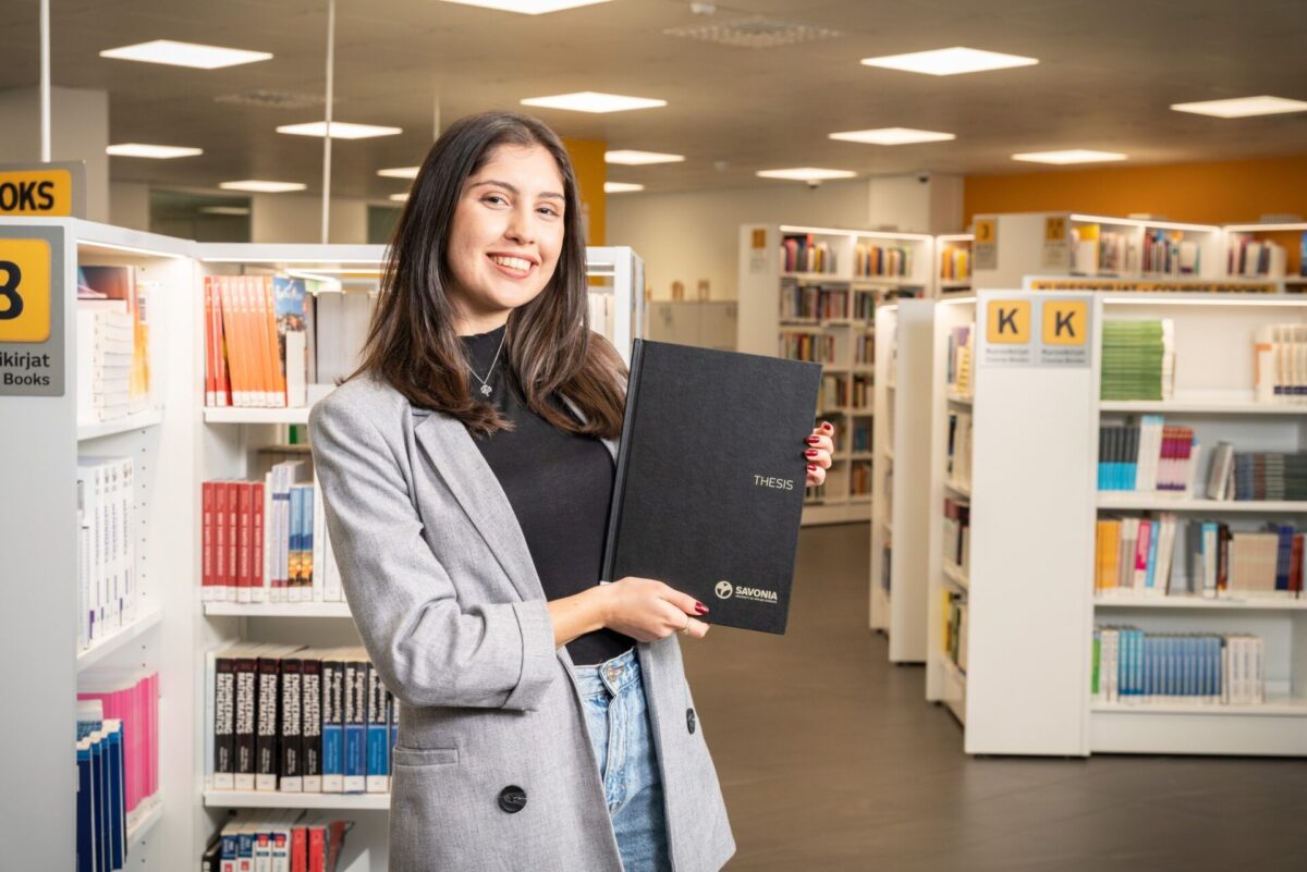Naisopiskelija kädessään opinnäytetyö kirjastossa