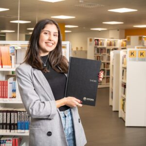 Naisopiskelija kädessään opinnäytetyö kirjastossa