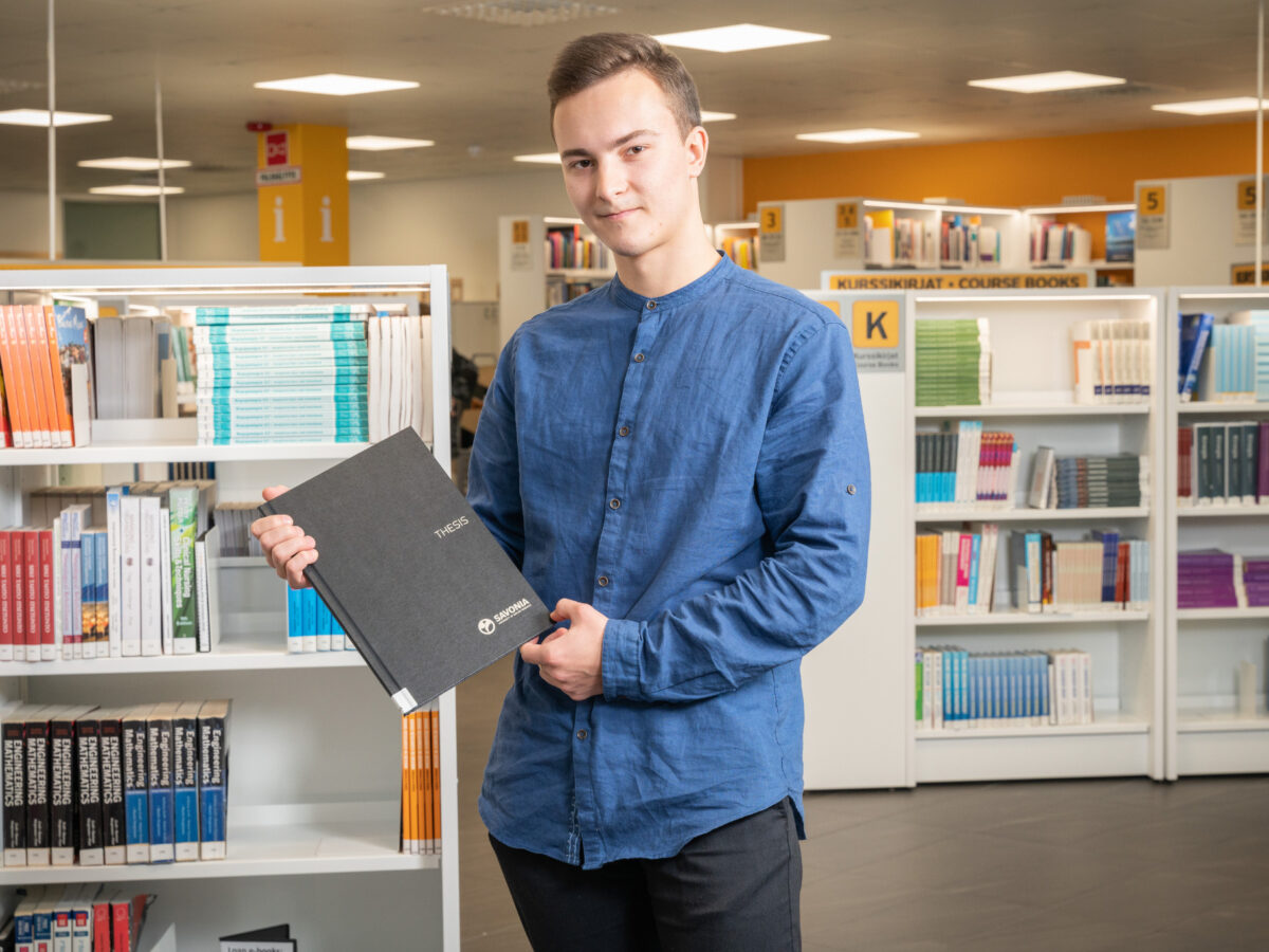 A student of Savonia standing up in the library and holding a thesis in his hands