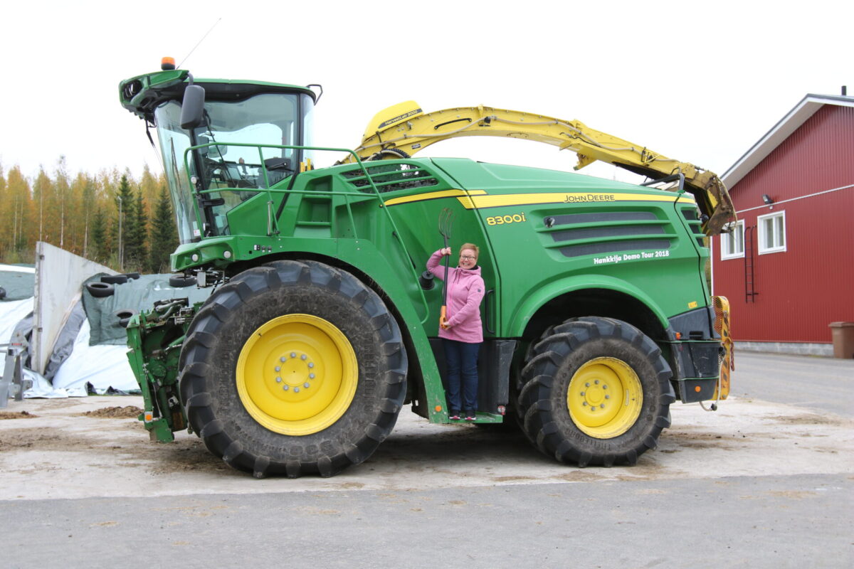 Iso vihreä traktori, jonka vieressä poseeraa hymyilevä nainen.