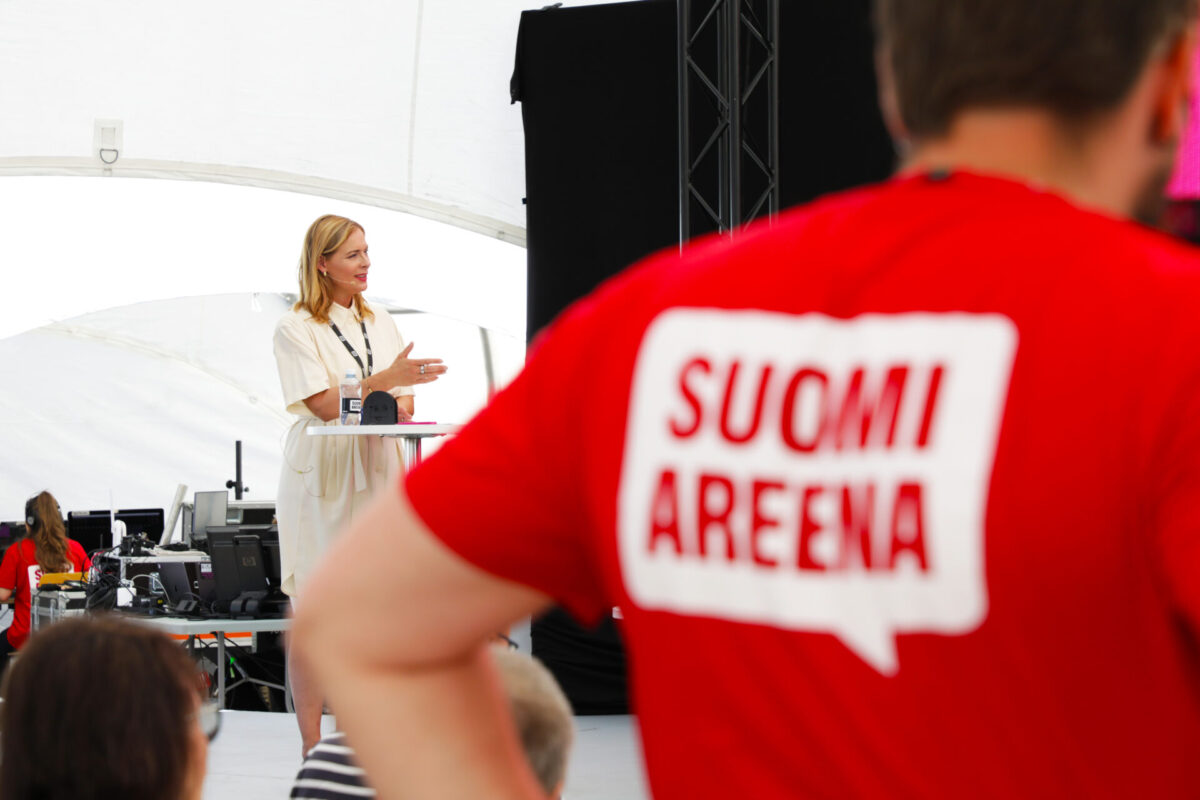 Etualalla t-paita, jossa lukee SuomiAreena, taka-alalla nainen esiintymislavalla.