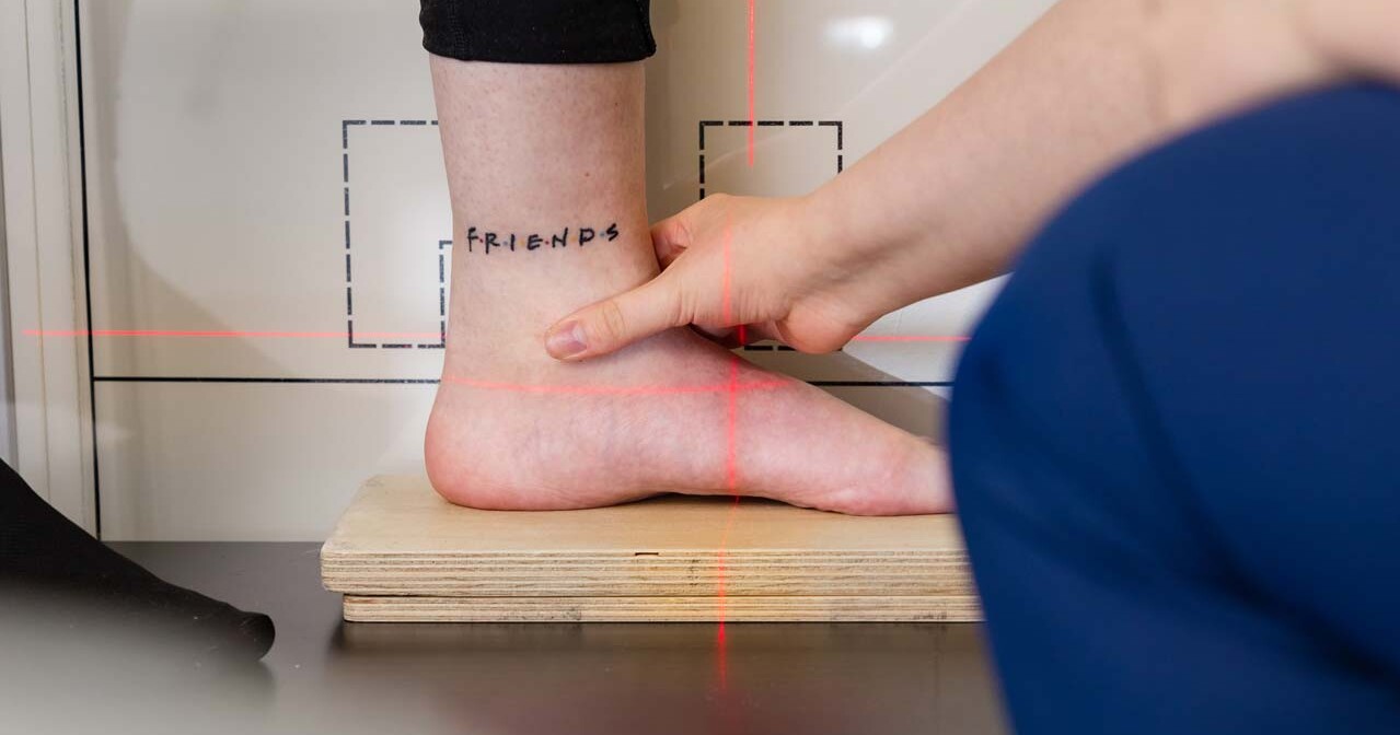 Henkilön nilkka, jossa on FRIENDS-tatuointi isoilla kirjaimilla, on toisen henkilön hellässä otteessa. Jalka on asetettu puulautaan, ja taustalla on punaisia laserviivoja ja -merkintöjä.