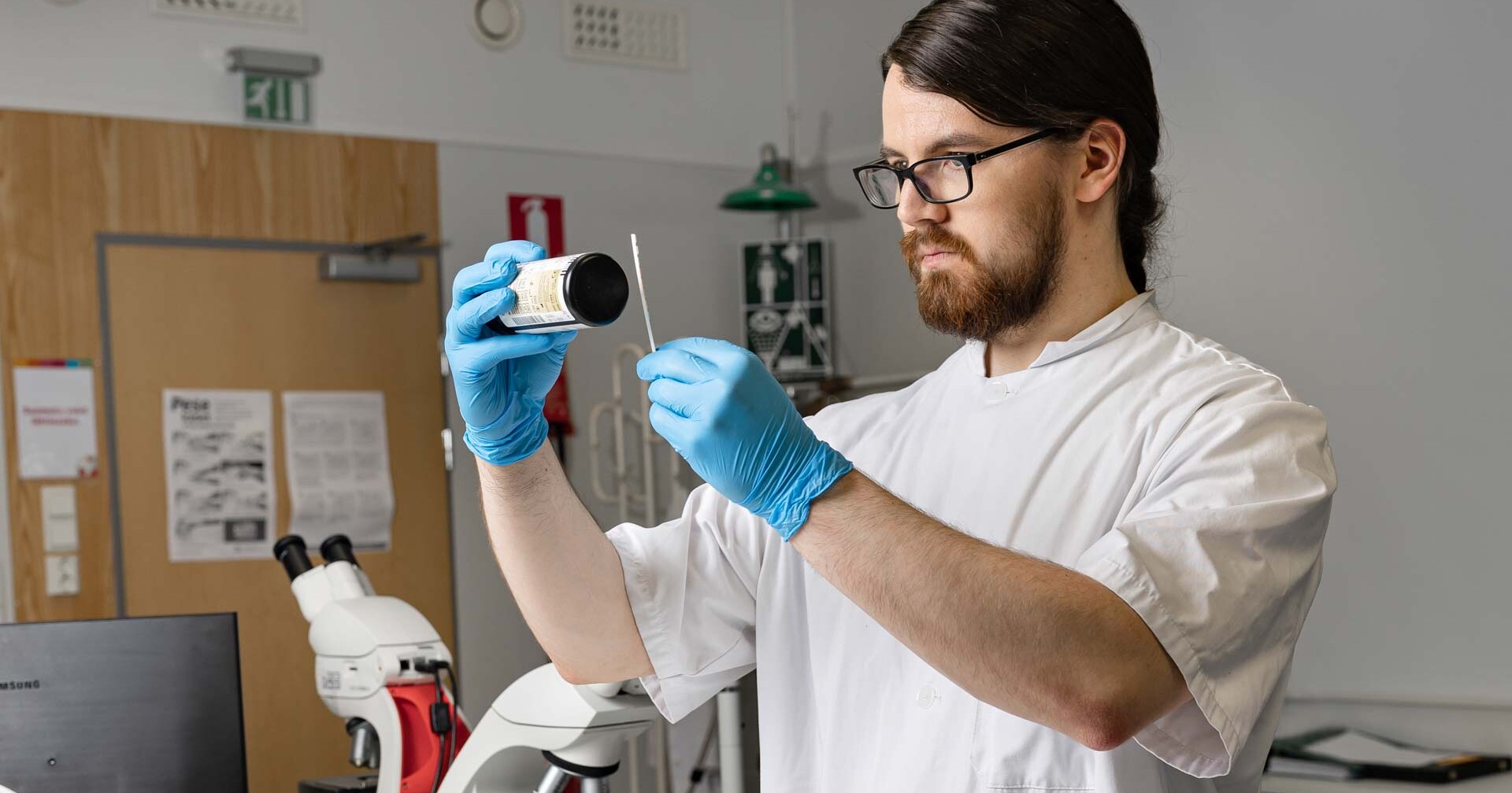 Laboratoriotakkiin ja sinisiin hanskoihin pukeutunut mies tutkii näyteastiaa ja pyyhkäisyä laboratoriossa, taustalla näkyy tieteellisiä laitteita ja asiakirjoja.