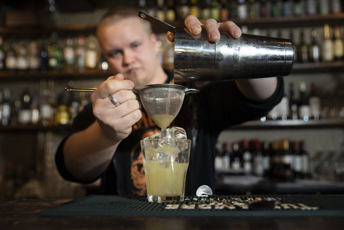 Henkilö kaataa baaristiksillä keltaista drinkkiä lasiin shakerista.