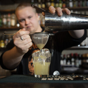 Henkilö kaataa baaristiksillä keltaista drinkkiä lasiin shakerista.