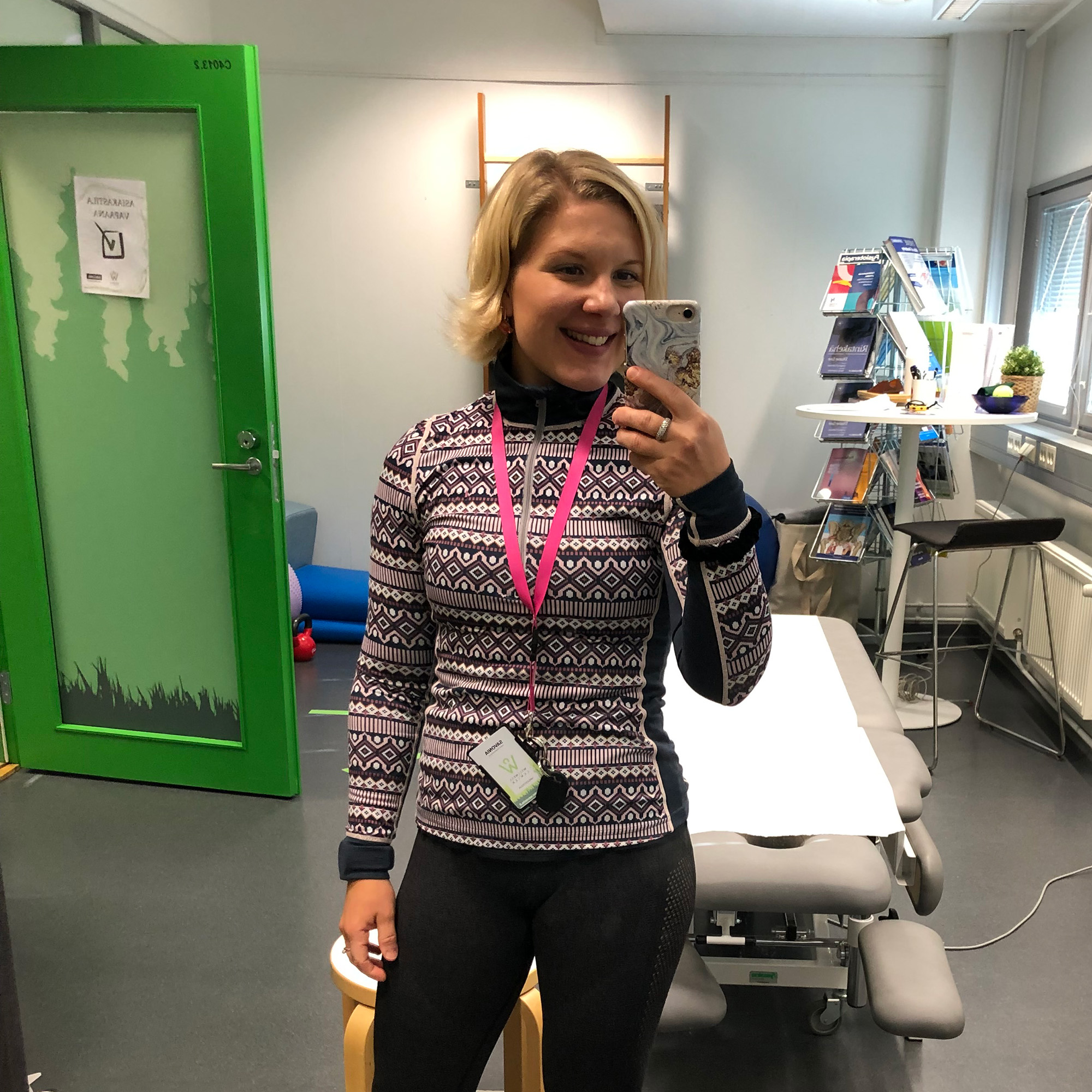 Hymyilevä henkilö, jolla on lyhyet vaaleat hiukset, ottaa peiliselfien valoisassa huoneessa, yllään kuviollinen pitkähihainen toppi, lantiopusero ja tummat housut. Takana on vihreä ovi, terapiapöytiä ja hyllyjä, joissa on lehtiä.