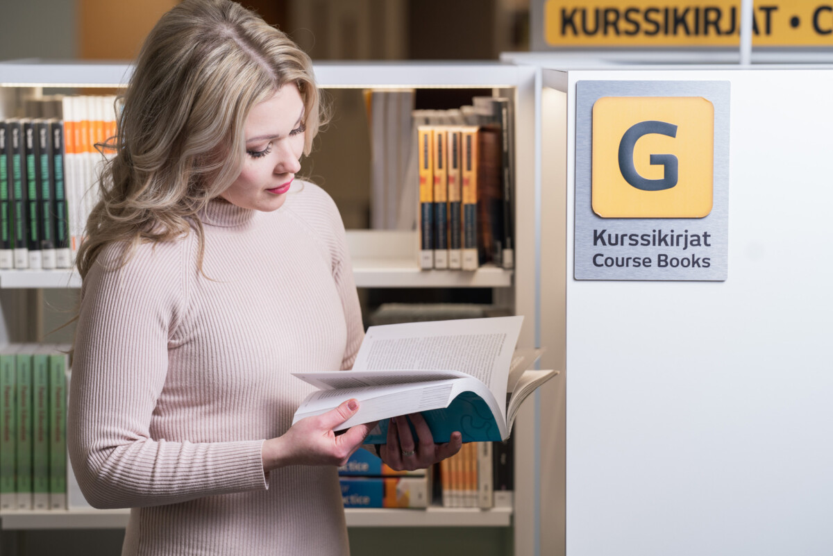 Opiskelija seisoo kirjastossa kirja kädessä.