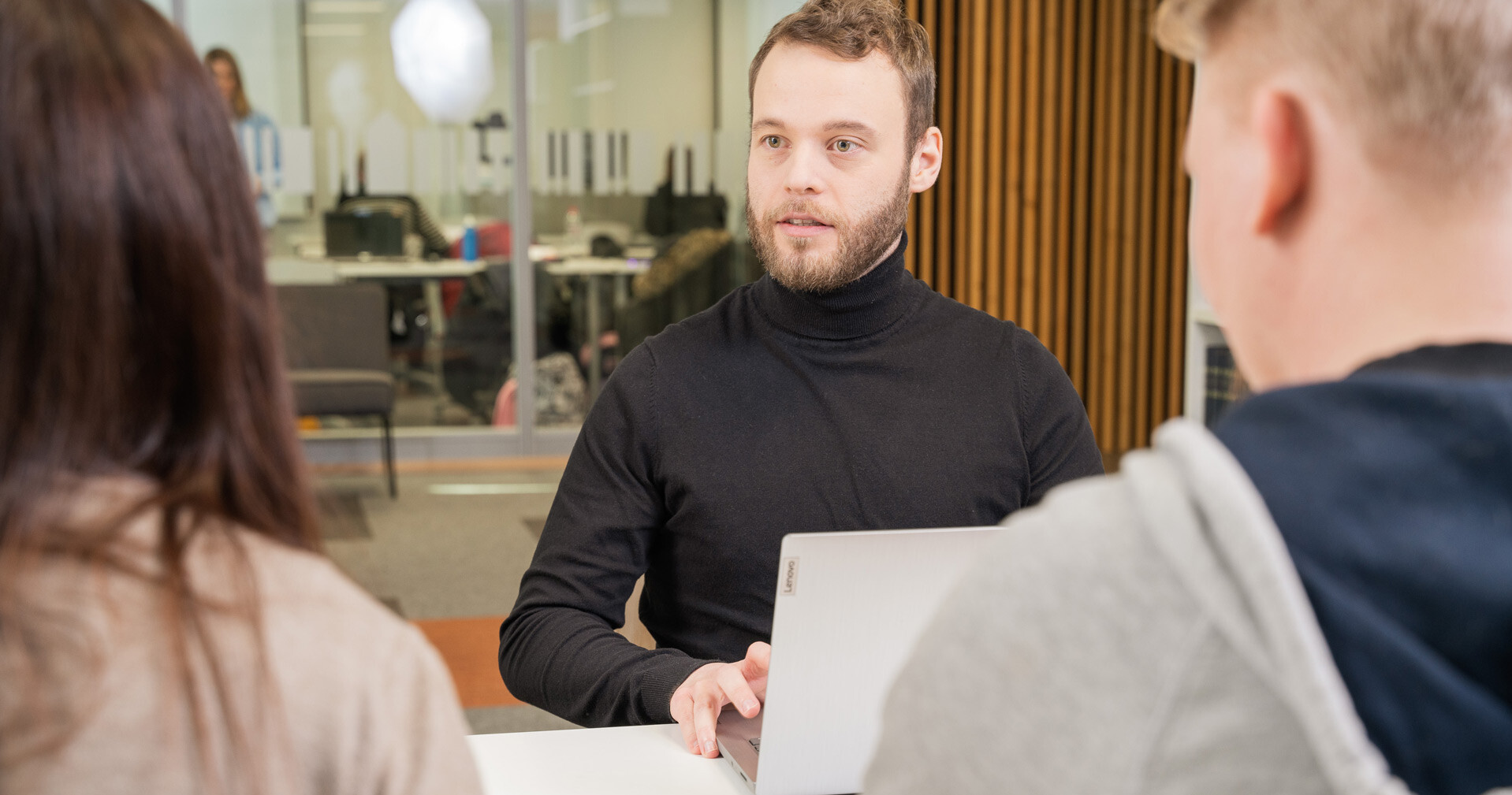 Parrakas mies, jolla on musta poolokaulus, istuu pöydän ääressä kannettavan tietokoneen kanssa ja keskustelee kahden henkilön kanssa, joiden selkä on kameraan päin, modernissa toimistoympäristössä.