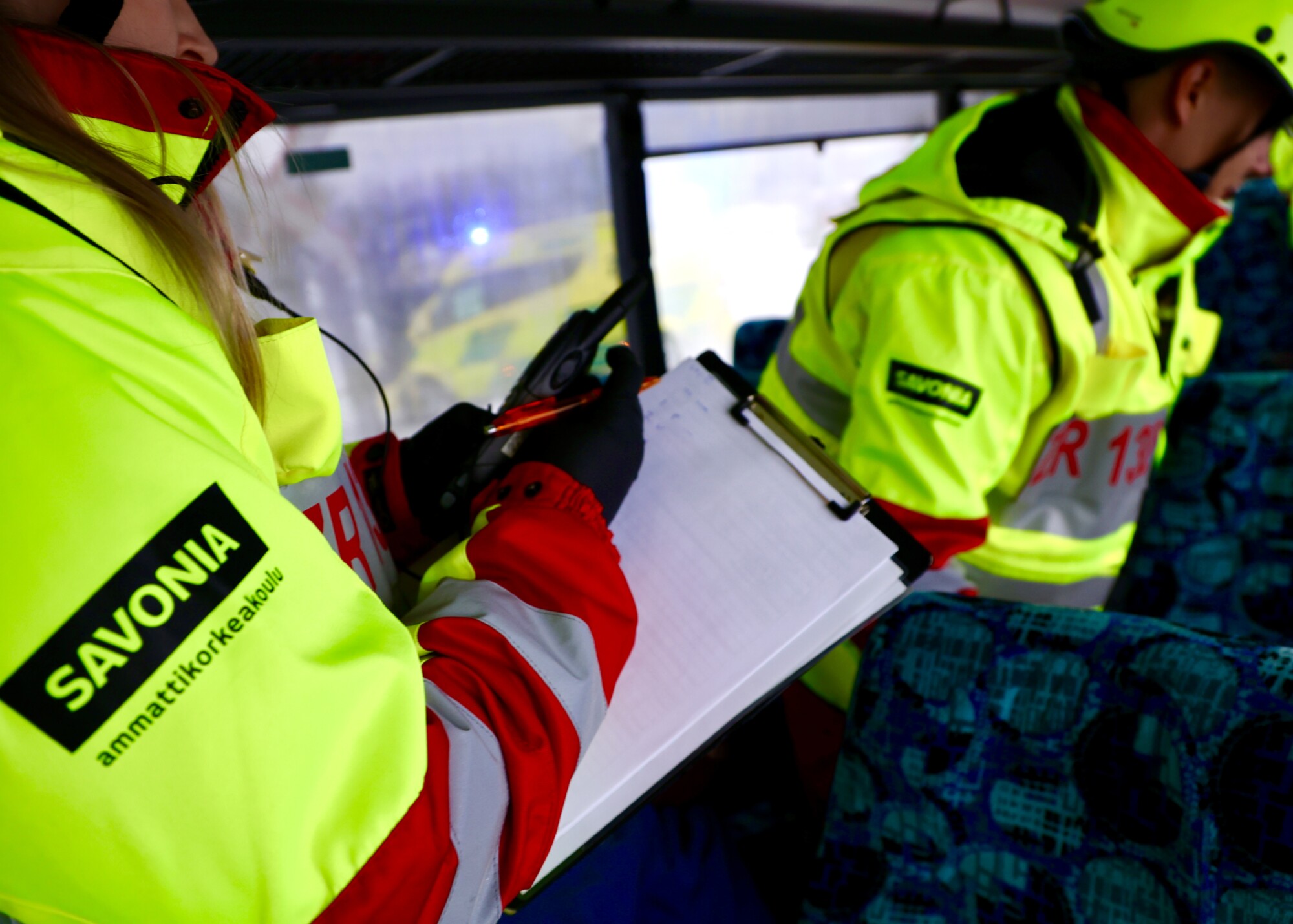 Lähikuvassa näkyy osa ensihoitajaa, joka kirjoittaa muistiinpanoja, taustalla toinen ensihoitaja.