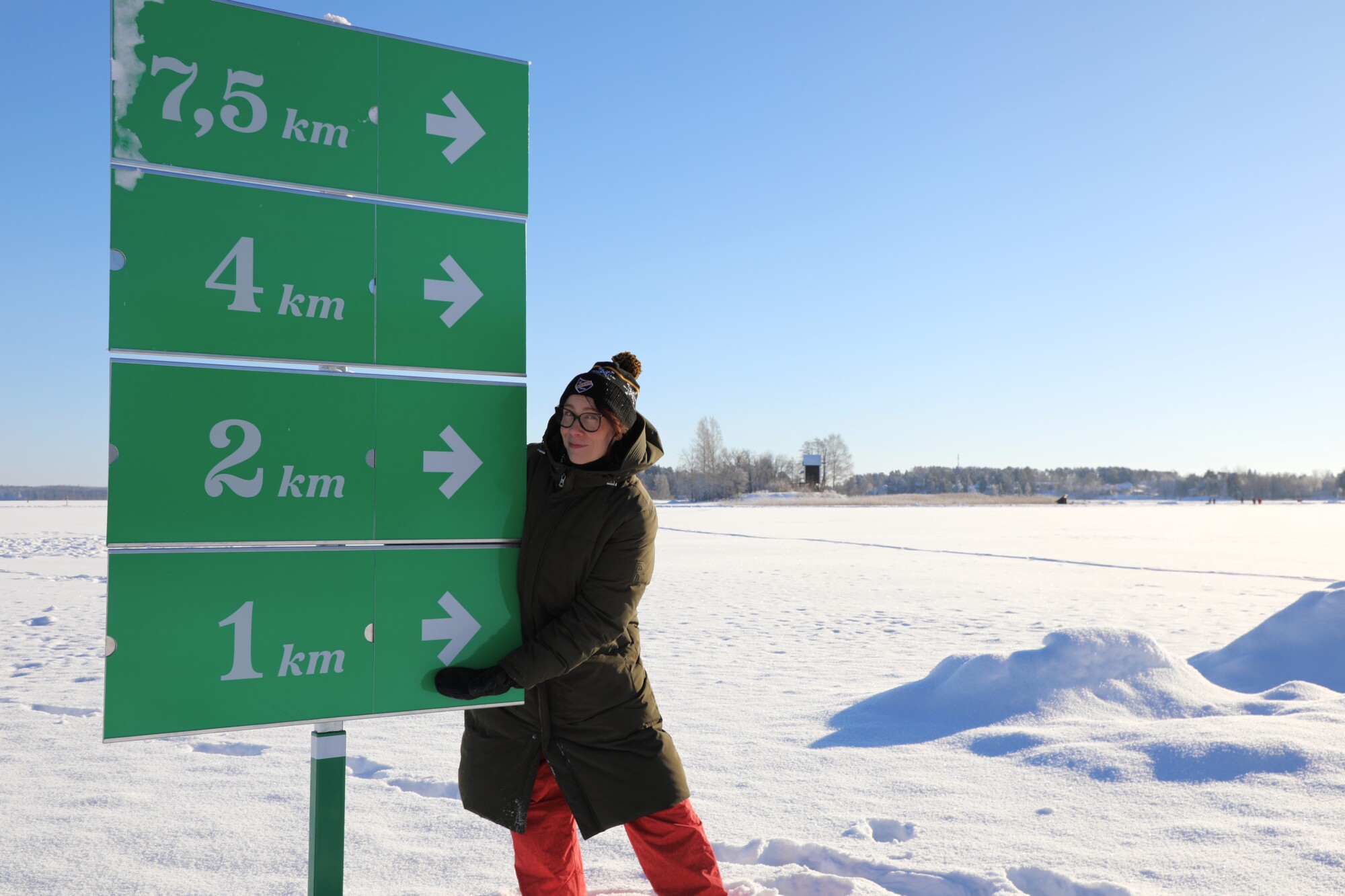 Miia Maaninen seisoo talvisella Kallaveden jäällä aurinkoisessa säässä ja nojaa kilometrikylttiin.