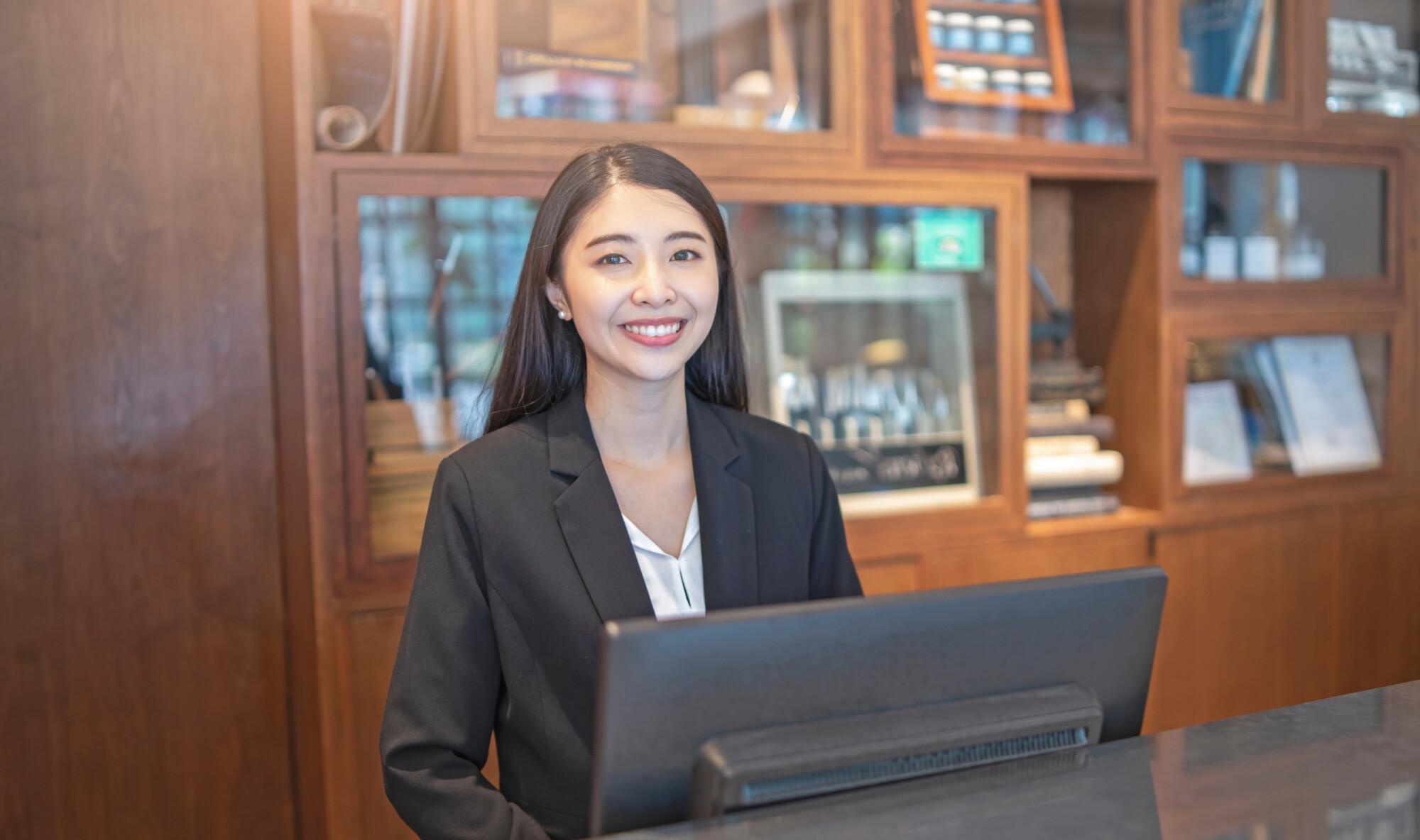 asian woman hotel receptionist standing behind a marble counter in front of a computer monitor smiling, long black hair wearing a suit and white shirt with wooden cabinets and books at the background.