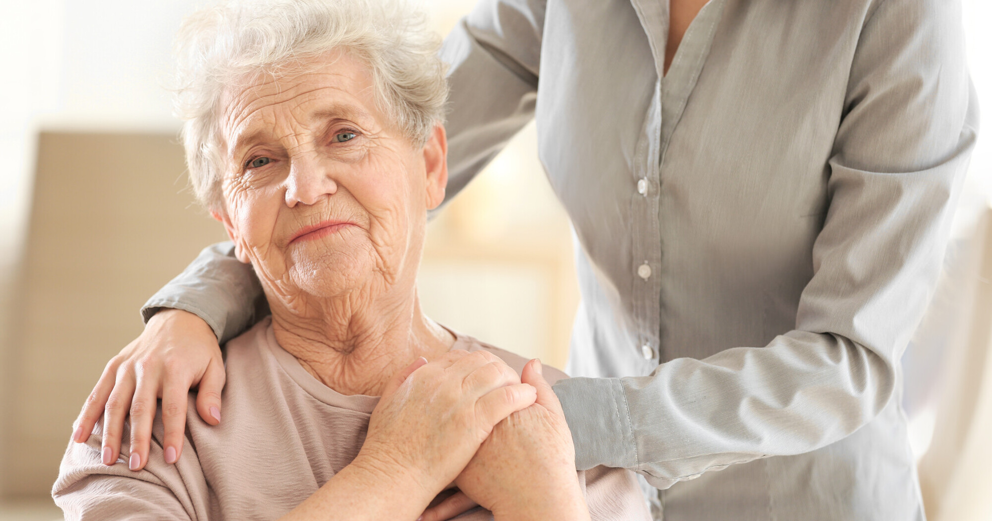 Vanha nainen, taustalla näkyy ihmisen kädet hänen olkapäillään ja käsi kädessä.