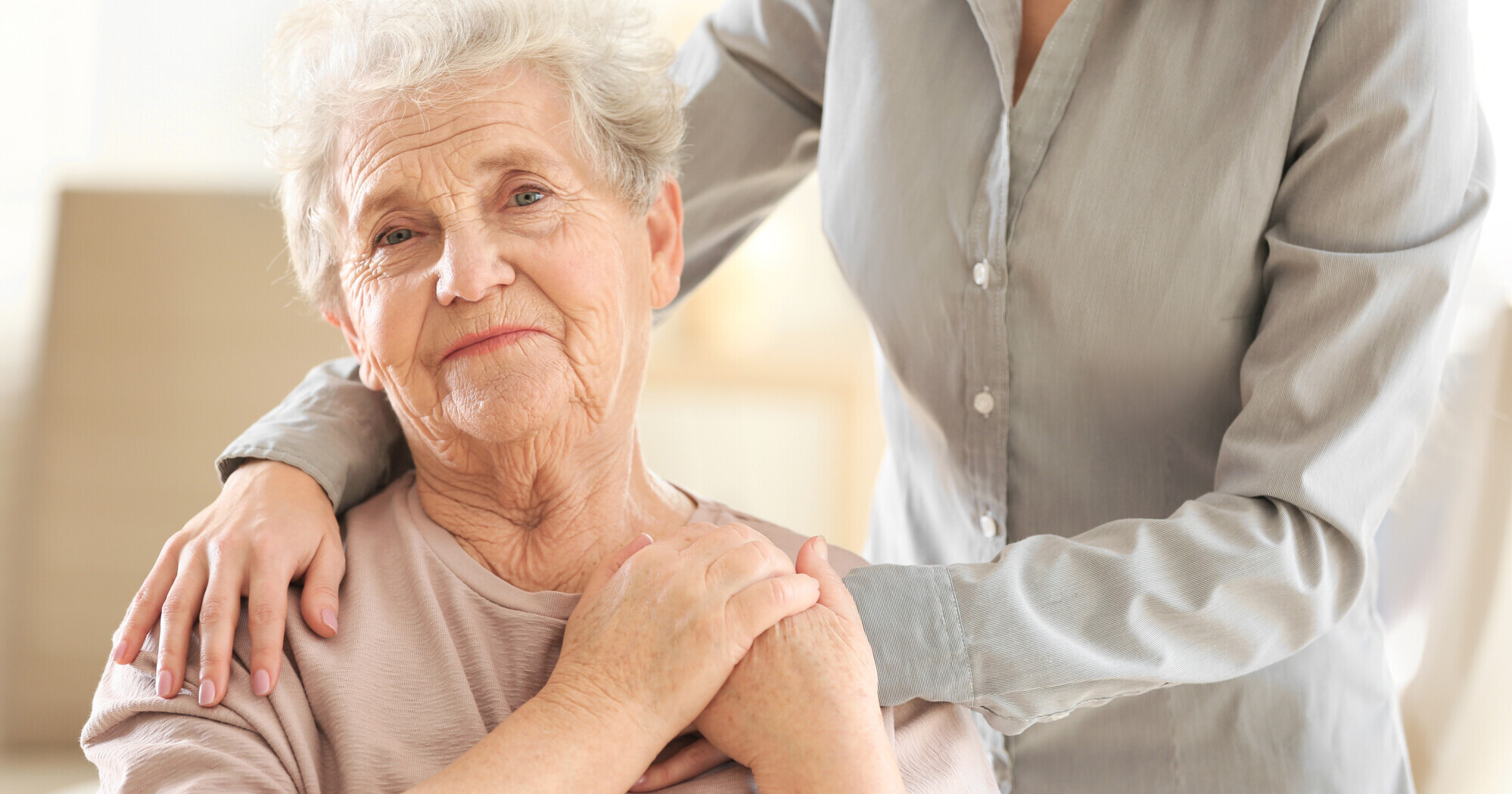Vanha nainen, taustalla näkyy ihmisen kädet hänen olkapäillään ja käsi kädessä.