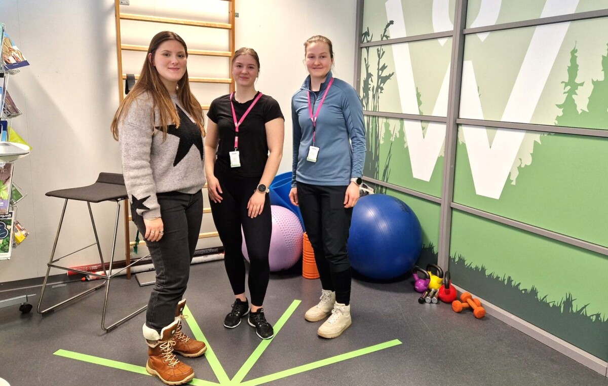 Kolme naista seisoo vierekkäin fysioterapiatiloissa, takan jumppapalloja, käsipainoja ja muuta välineistöä.