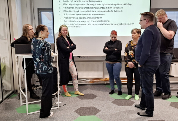 Ihmisiä seisomassa lattialla, jossa värikkäitä kuvioita, taustalla valkokankaalla tekstiä.