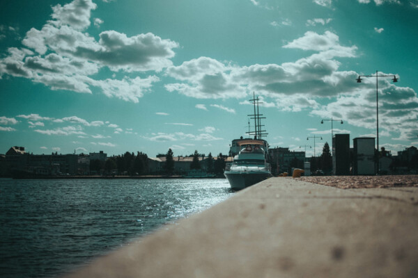 Jahti on telakoituna rantaviivaan pilvisen, sinivihreän taivaan alla. Rakennukset ja katuvalot reunustavat kaukaista rantaa, ja auringonvalo heijastuu vedestä.