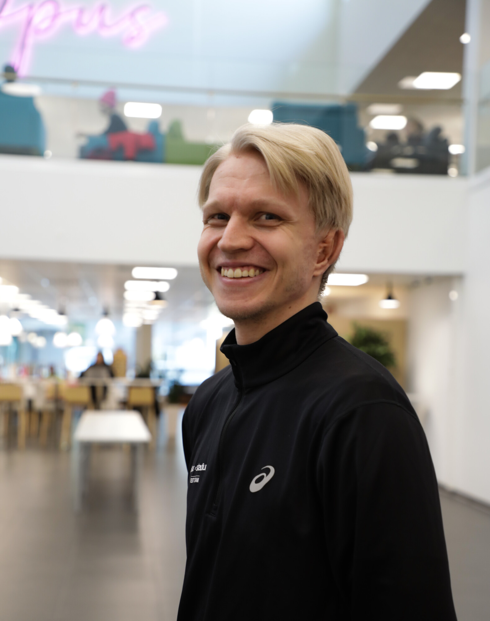Hymyilevä vaaleatukkainen mies, jolla on yllään musta Asics-takki, seisoo sisätiloissa modernissa, avoimessa tilassa, jossa on pöytiä ja ihmisiä taustalla.