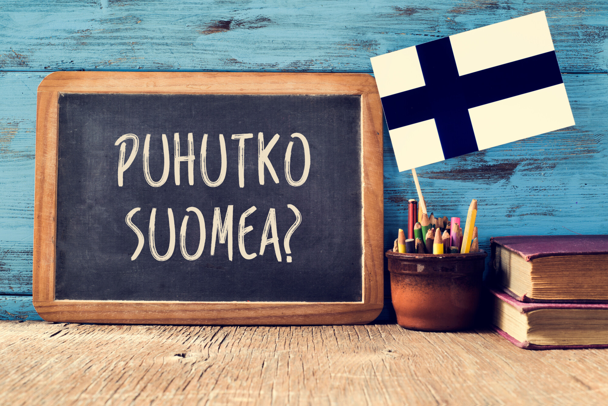 Liitutaulu, jossa lukee Puhutko suomea?, pienen Suomen lipun vieressä värikynien purkissa ja kaksi pinottua kirjaa puupöydällä, taustalla sininen puinen seinä.