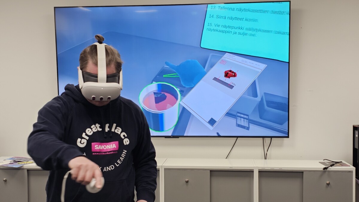 Virtuaalitodellisuuslaseja käyttävä ja ohjainta pitelevä henkilö on vuorovaikutuksessa VR-ympäristön kanssa, joka näkyy hänen takanaan suurella näytöllä, jossa on 3D-objekteja ja suomenkielistä tekstiä.