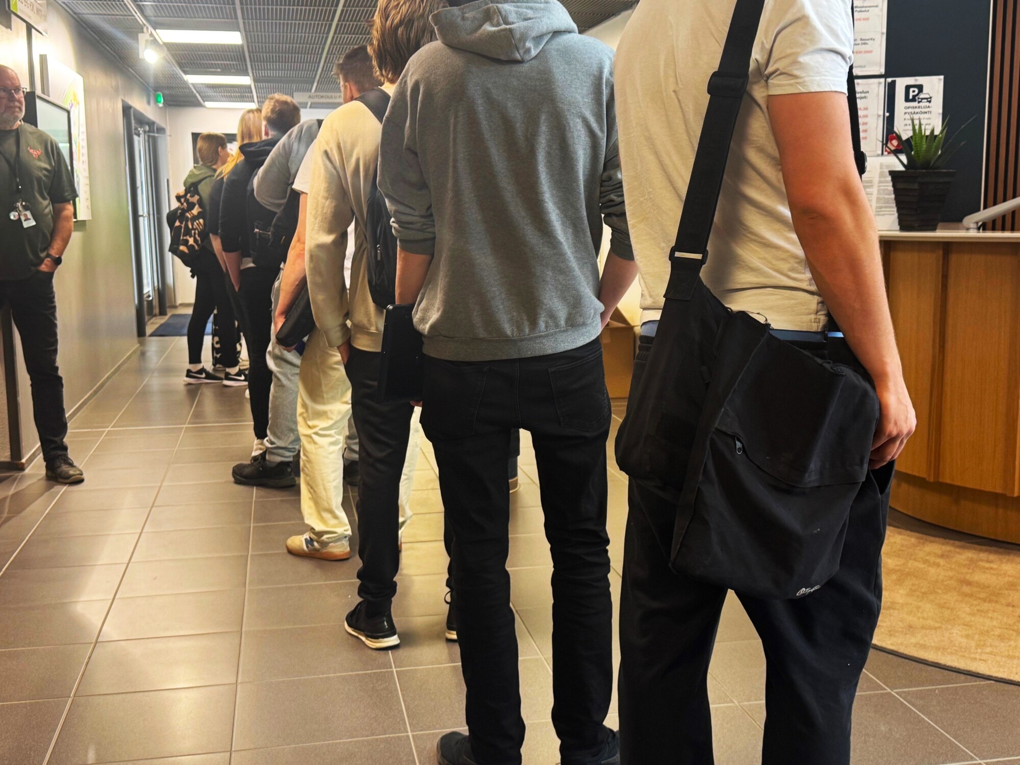 Ihmiset seisovat jonossa rakennuksen sisällä odottamassa vuoroaan. Useimmat ovat pukeutuneet rennosti ja kantavat laukkuja. Miljöö näyttää olevan vastaanotto- tai palvelupistealue, jossa on laattalattia ja moderni sisustus.