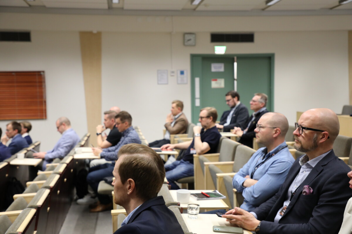 Ryhmä miehiä istuu riveissä luentosalissa ja kuuntelee tarkkaavaisesti esitystä. Jotkut tekevät muistiinpanoja, ja muutamilla on edessään kannettava tietokone tai muistikirja. Ympäristö vaikuttaa muodolliselta ja ammattimaiselta.