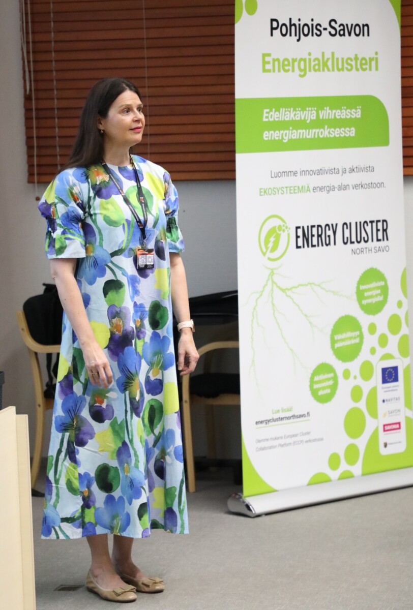 Siniseen kukkamekkoon pukeutunut nainen seisoo Pohjois-Savonin energiaklusterin banderollin lähellä. Banderollissa on vihreää tekstiä ja logoja, jotka liittyvät energiainnovaatioihin ja kestävään kehitykseen.
