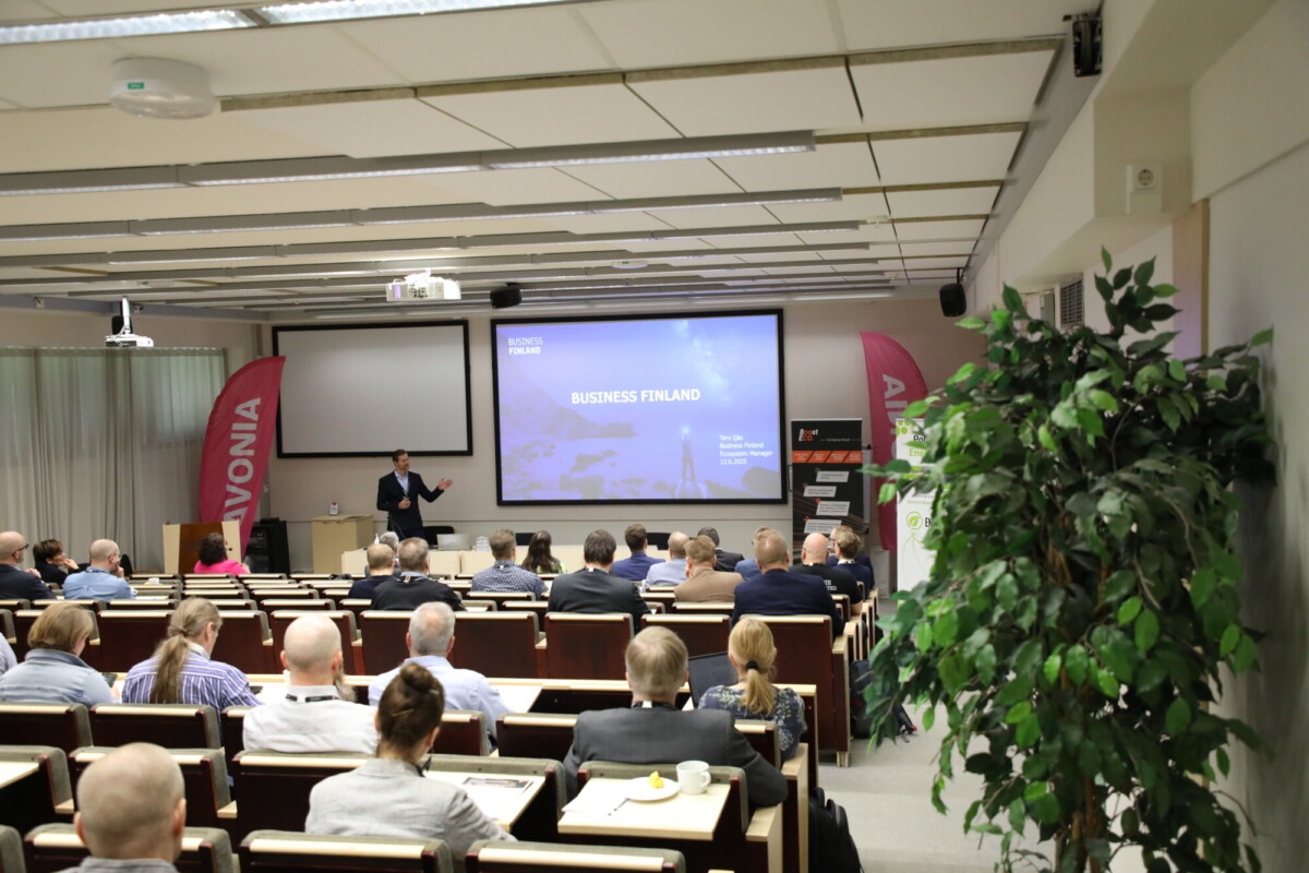 Mies pitää esityksen nimeltä Business Finland yleisölle modernissa luentosalissa, jossa on rivit työpöytiä, suuri valkokangas ja bannereita molemmin puolin. Etualalla näkyy kasvi.