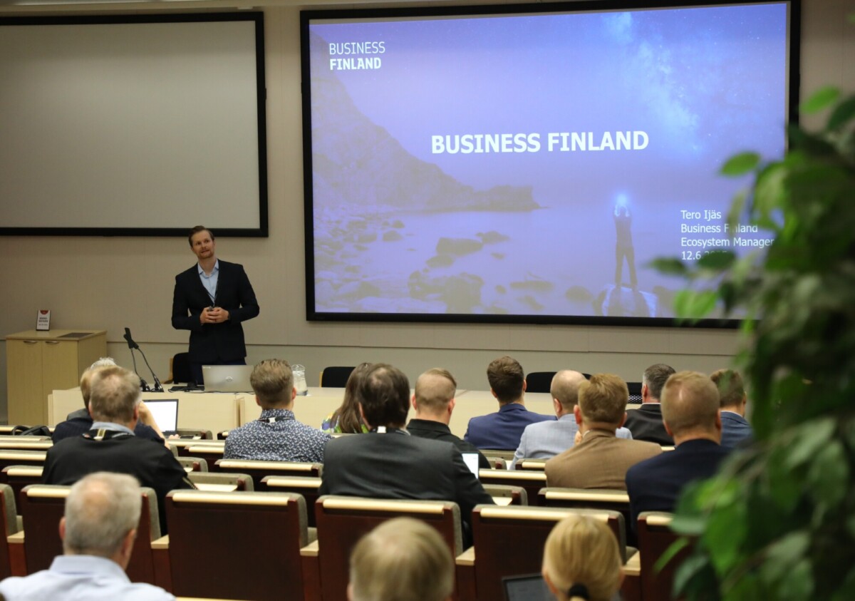 Mies seisoo puhujakorokkeella pitämässä esitystä yleisölle luentosalissa. Hänen takanaan olevalla suurella näytöllä näkyy Business Finlandin tiedot ja esityksen tiedot. Ihmiset istuvat ja kuuntelevat tarkkaavaisesti.