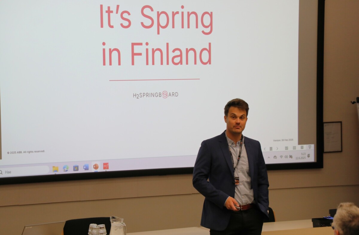 Siniseen bleiseriin pukeutunut mies seisoo heijastetun dian edessä, jossa lukee Kevät Suomessa. Huoneessa on pöytä lasikannuilla ja kannettava tietokone.