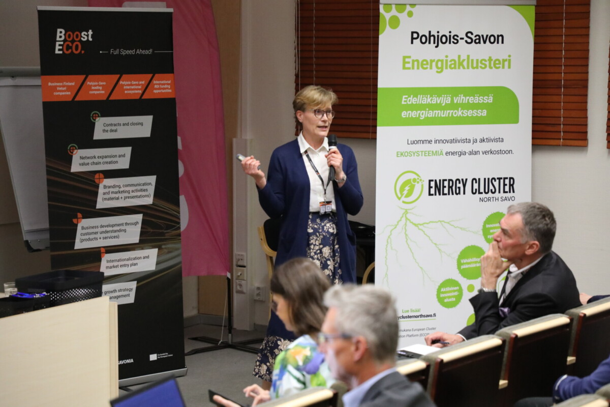 Nainen puhuu mikrofonin kanssa kokoushuoneessa. Hänen takanaan on bannereita, joissa on tekstiä energiasta, innovaatioista ja ympäristöystävällisistä aloitteista. Yleisö istuu ja kuuntelee tarkkaavaisesti.