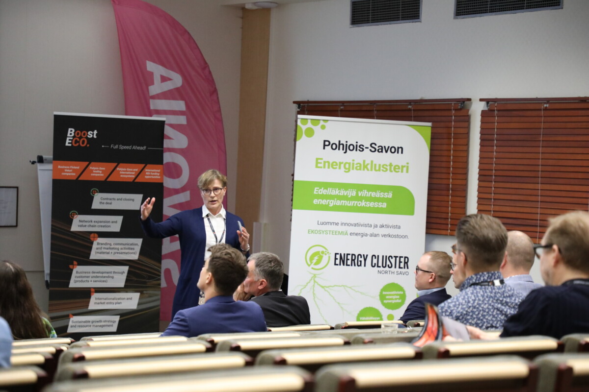 Bisnesasuihin pukeutunut henkilö puhuu yleisölle luentosalissa elehtien yhdellä kädellä. Heidän takanaan on suomenkielisiä bannereita Energiaklusterista ja Boost Ecosta. Läsnäolijat istuvat ja kuuntelevat tarkkaavaisesti.