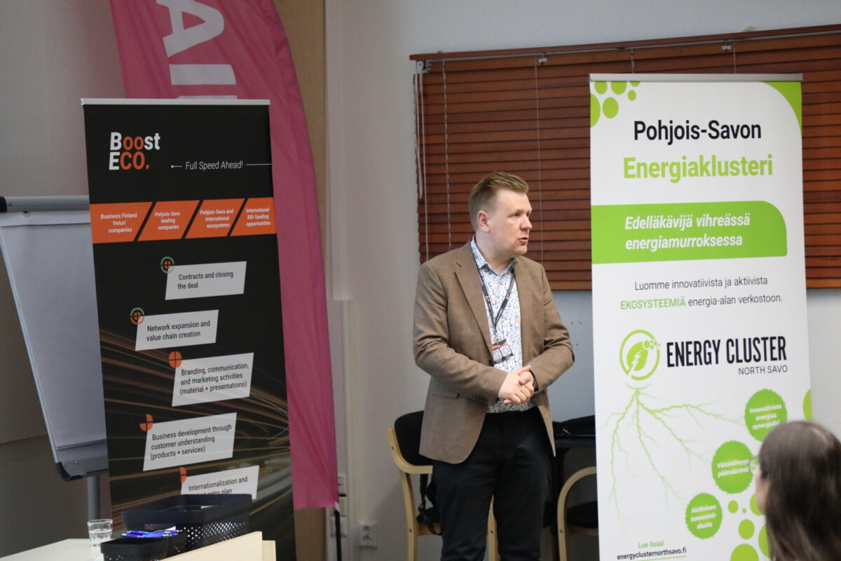 Pukuun pukeutunut mies seisoo kahden esittelybannerin edessä sisätilaisuudessa. Toinen banneri on Boost ECOlle ja toinen Pohjois-Savon Energiaklusterille.