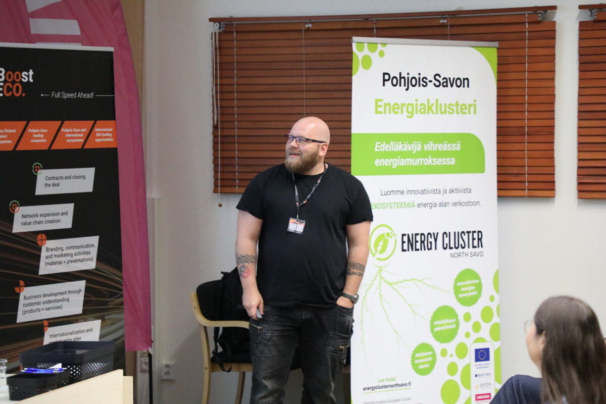 Kalju mies, jolla on silmälasit, parta ja tatuoinnit, seisoo puhumassa kahden pystysuoran bannerin edessä energiaryppäistä sisätilaisuudessa, ja yleisön istuimet näkyvät etualalla.